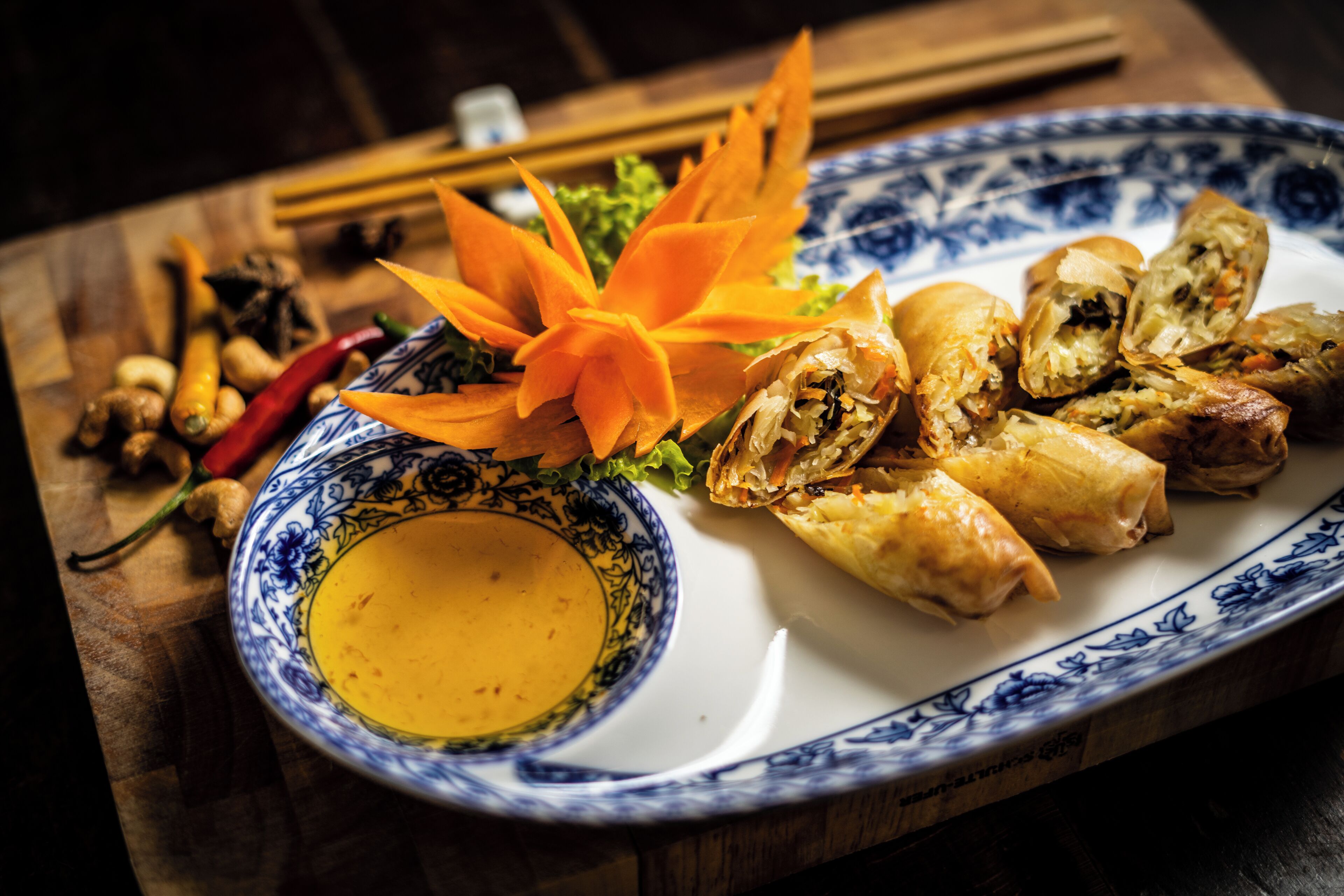 A plate of spring rolls with a decorative flower, a bowl of dipping sauce, and various vegetables on a wooden table.