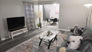 A living room with a flat-screen TV, a glass coffee table, a grey sofa, and a patterned rug.