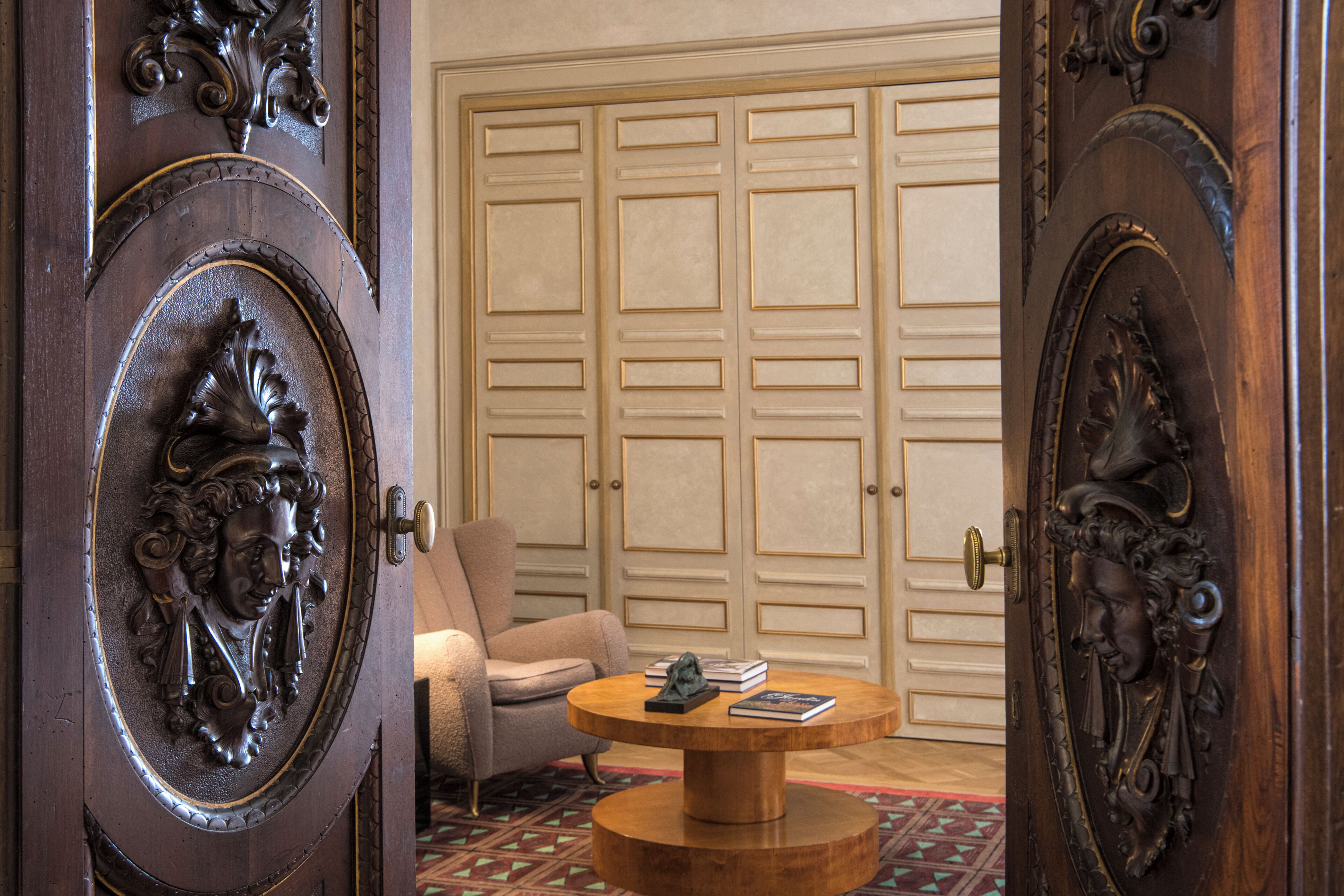 A room with a wooden door featuring intricate carvings, a round wooden coffee table with books, a beige armchair, and a large wooden closet in the background.