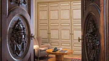 A room with a wooden door featuring intricate carvings, a round wooden coffee table with books, a beige armchair, and a large wooden closet in the background.
