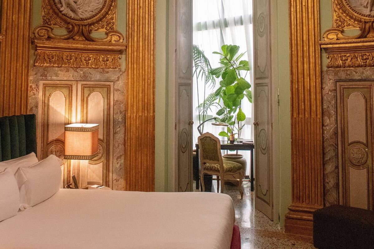 A luxurious hotel room with a large bed, ornate ceiling, and a view of a dining area with a plant.