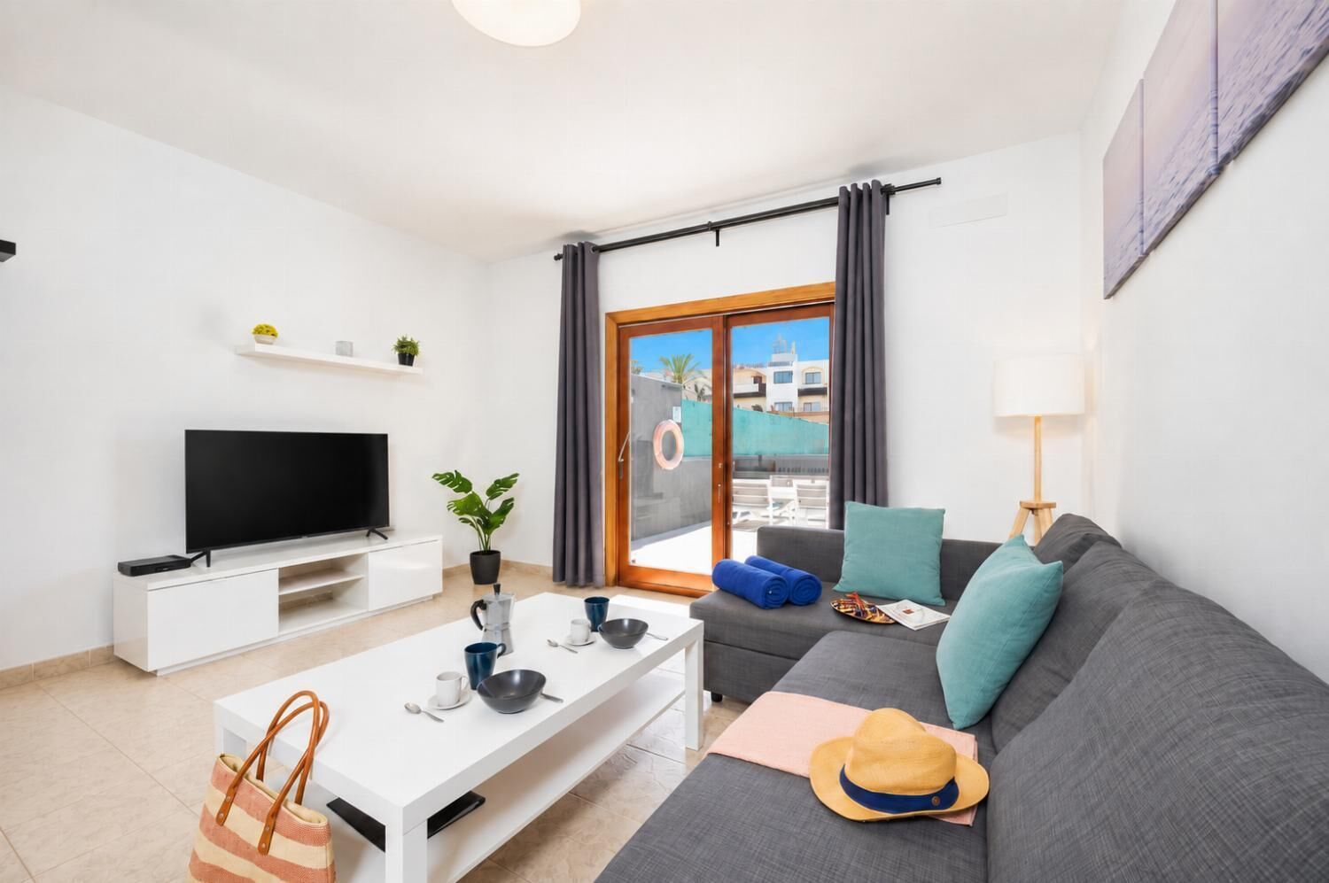 Ein modernes Wohnzimmer mit einem Flachbildfernseher, einem weißen Couchtisch, einem grauen Sofa und einem großen Fenster mit Vorhängen.
