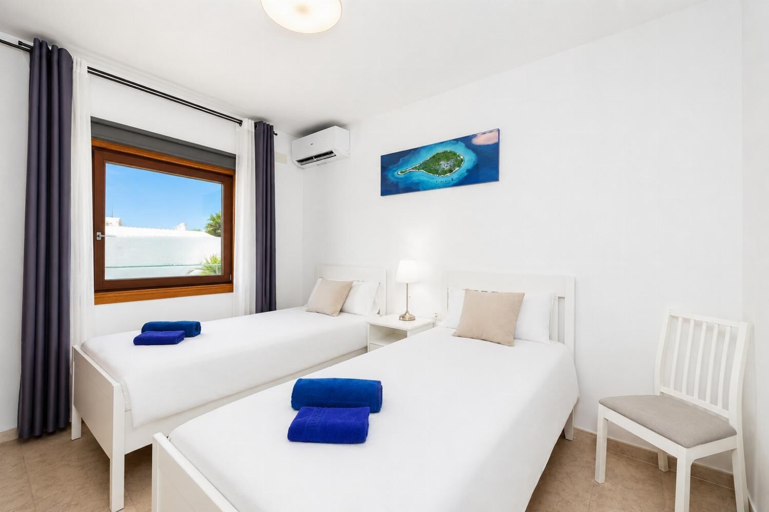 Habitación de hotel con dos camas, una ventana con vista al mar, una mesita con una lámpara y una silla.