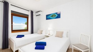 Quarto de hotel com duas camas, uma janela com vista para o mar, uma mesinha com abajur e uma cadeira.