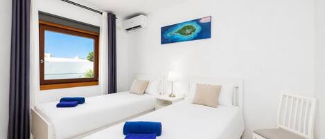 Habitación de hotel con dos camas, una ventana con vista al mar, una mesita con una lámpara y una silla.
