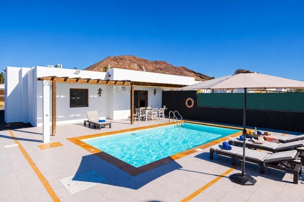 A swimming pool with lounge chairs and a covered area, surrounded by a white building with a mountain in the background.