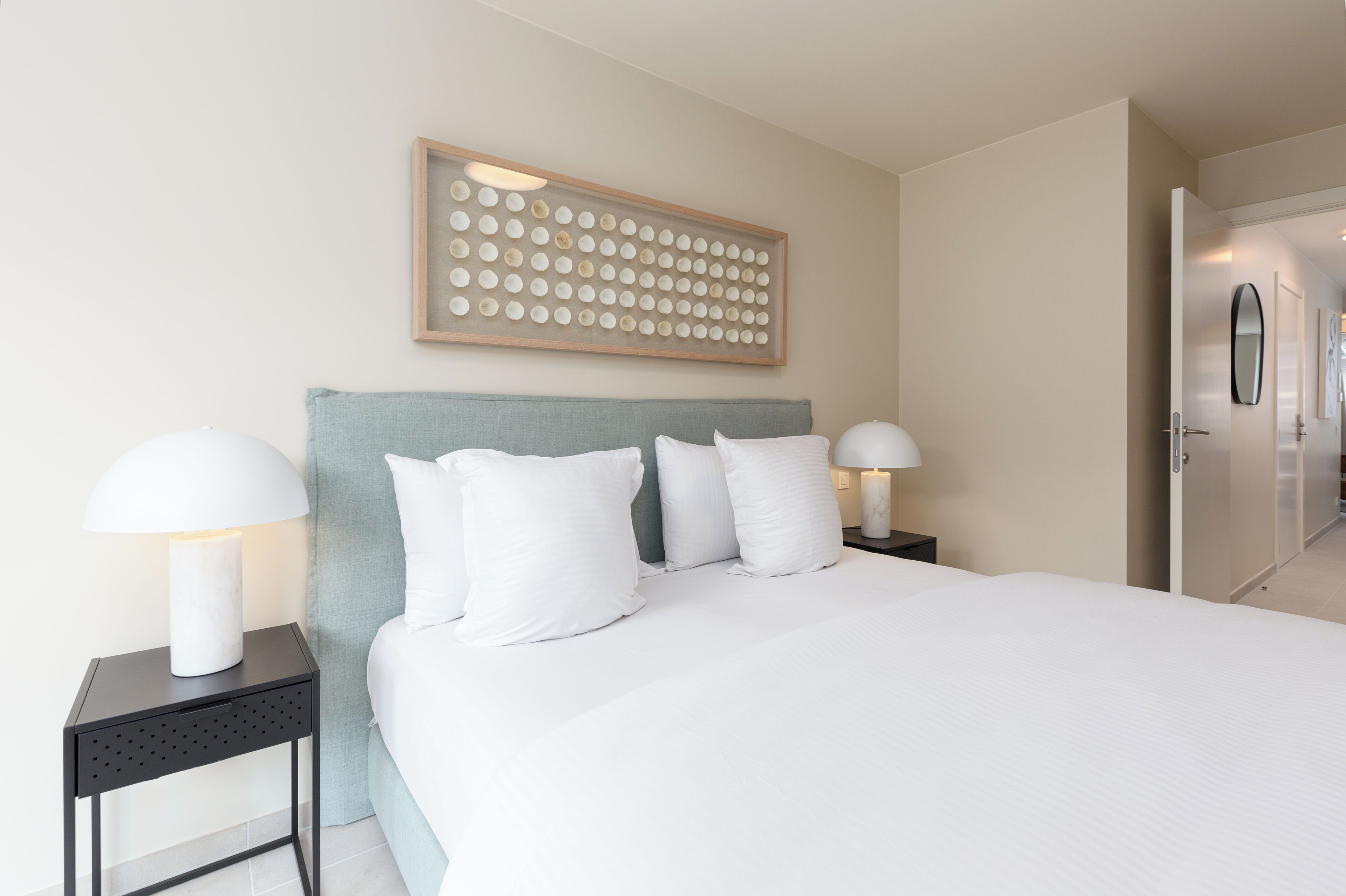 A modern hotel room with a large bed, two bedside tables with lamps, a wall-mounted artwork, and a door leading to another room.