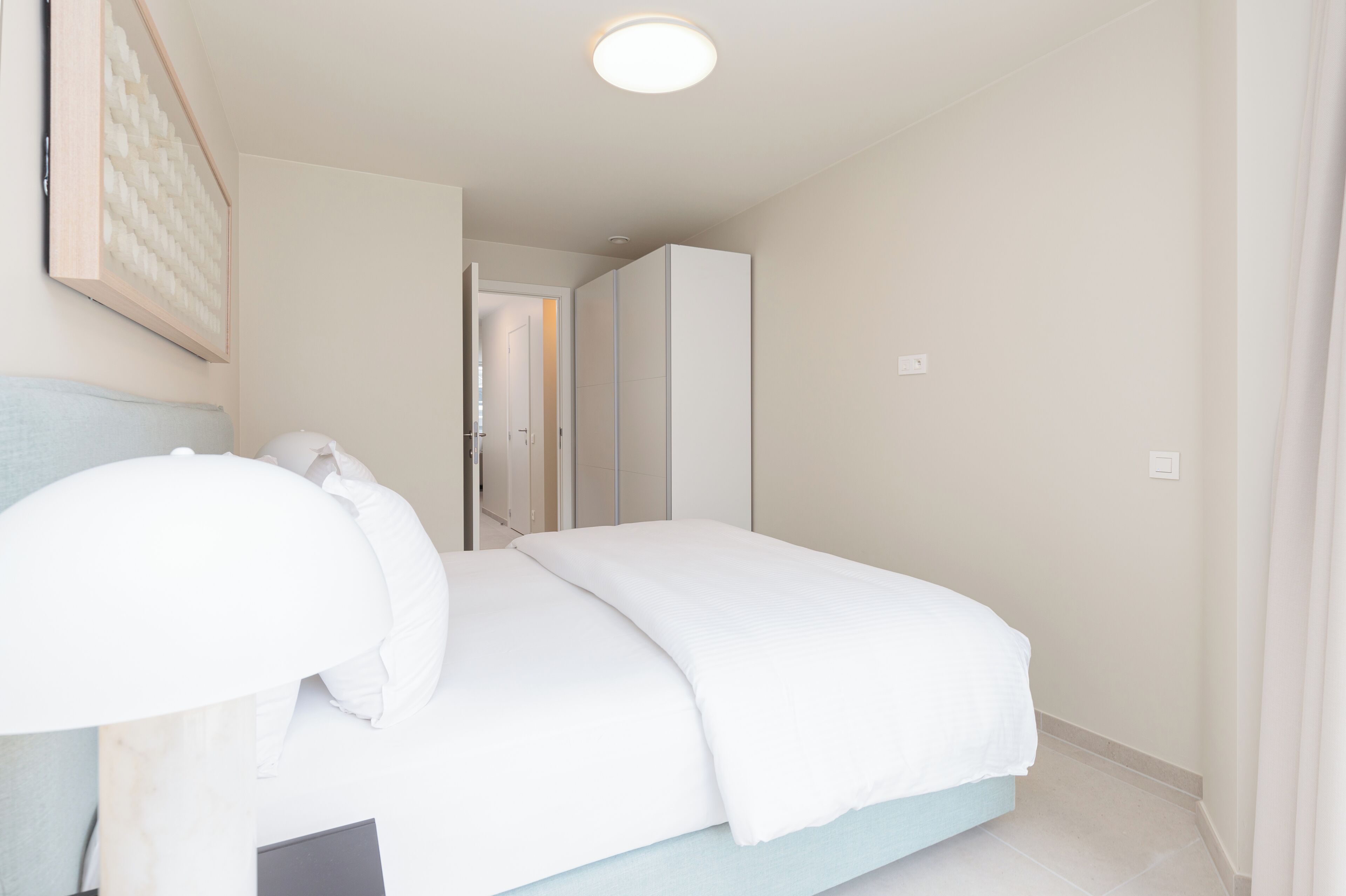 A bedroom with a bed, a bedside table, a lamp, and a door leading to another room.