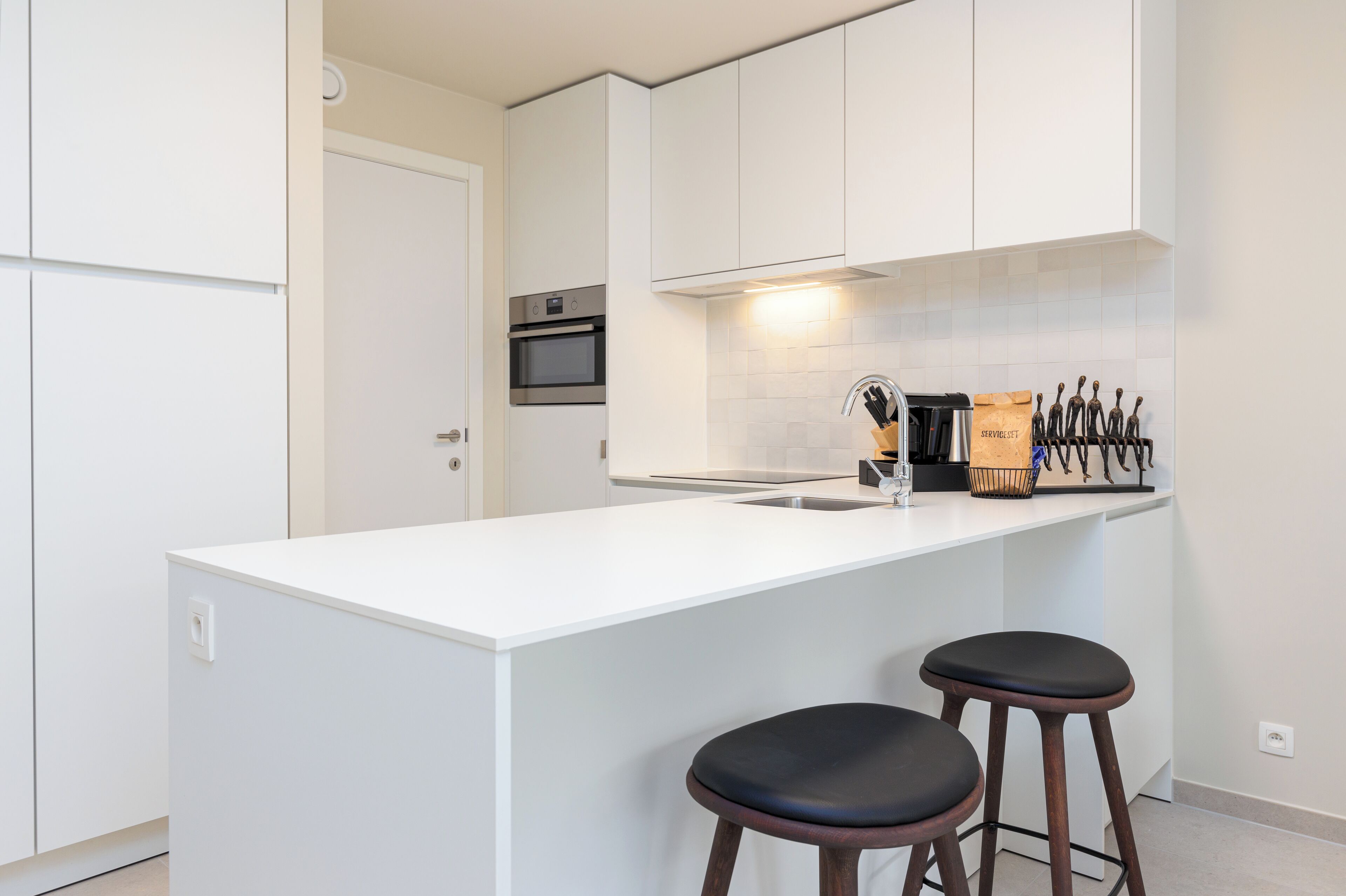 A modern kitchen with white cabinetry, a central island with two stools, a built-in oven, and a knife block on the wall.