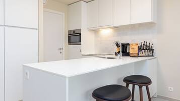 A modern kitchen with white cabinetry, a central island with two stools, a built-in oven, and a knife block on the wall.