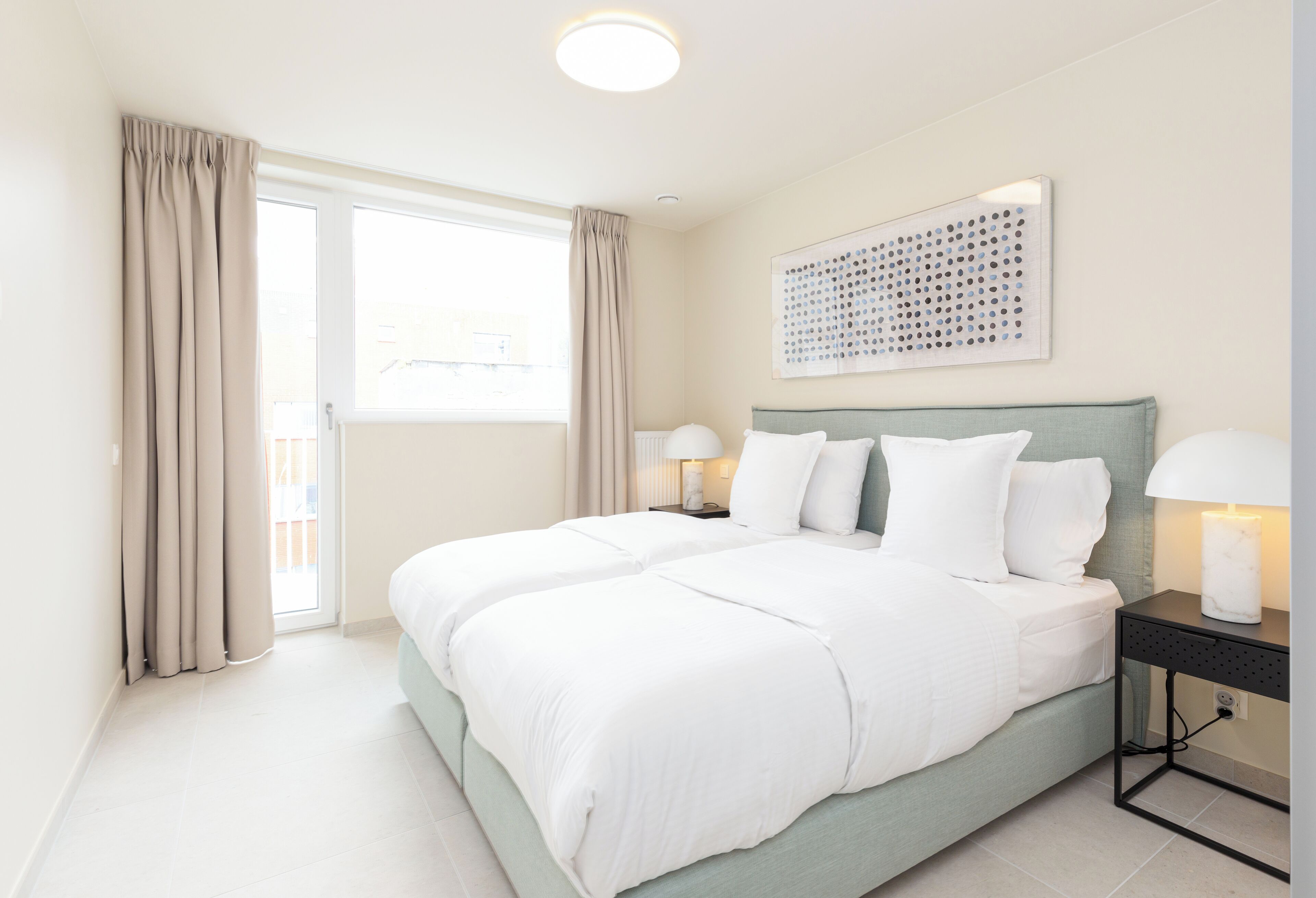 A modern bedroom with a large bed, a nightstand with lamps, a ceiling light, and a window with curtains.