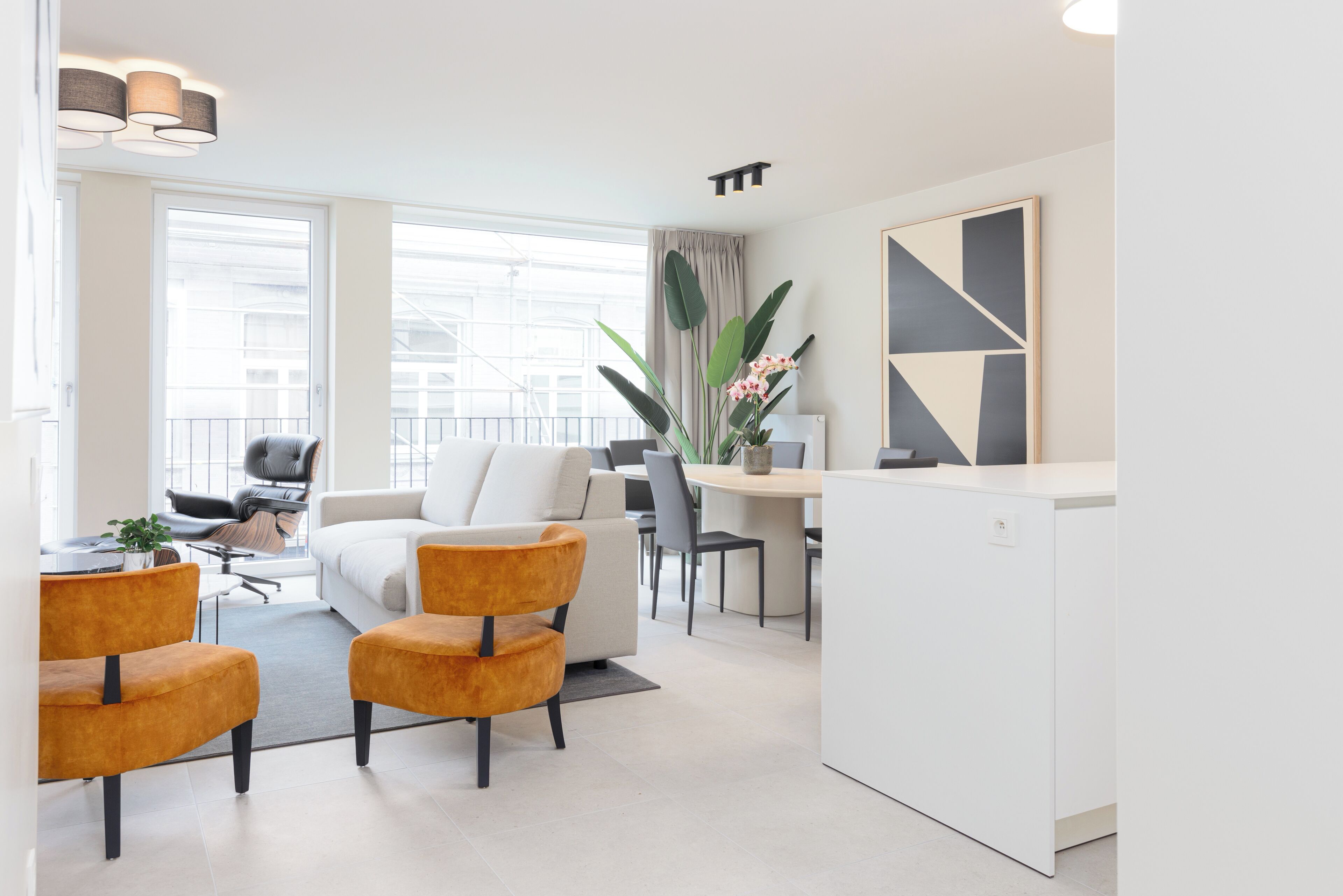 Een moderne woonkamer met een witte bank, twee oranje fauteuils, een eethoek met een tafel en stoelen, en een keuken met een witte kast.