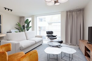 Uma sala de estar moderna com um sofá branco, poltronas laranja, uma mesa de centro e uma TV sobre um suporte de madeira.