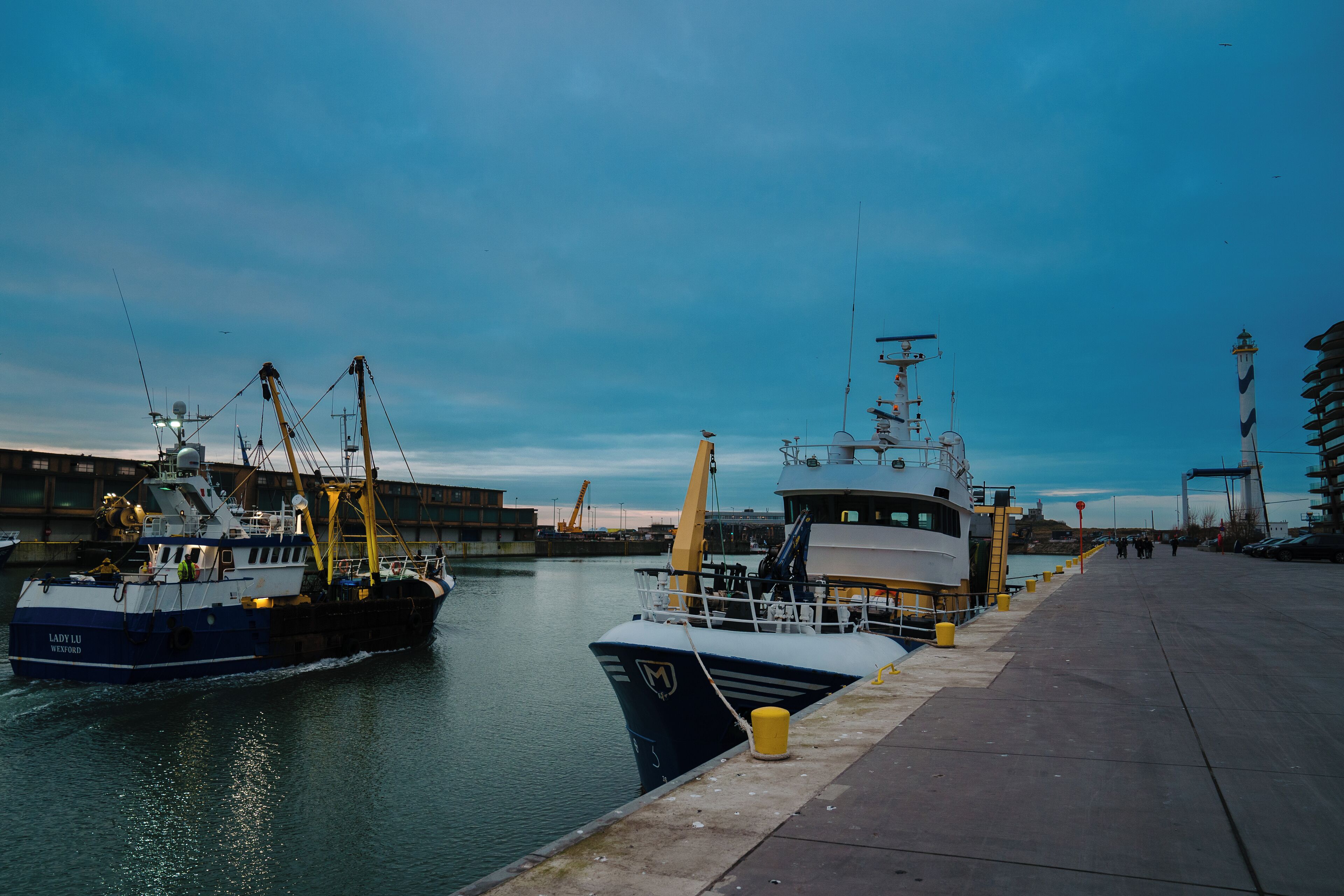A harbor with a large ship docked at a pier, a smaller boat, and a building with a crane.