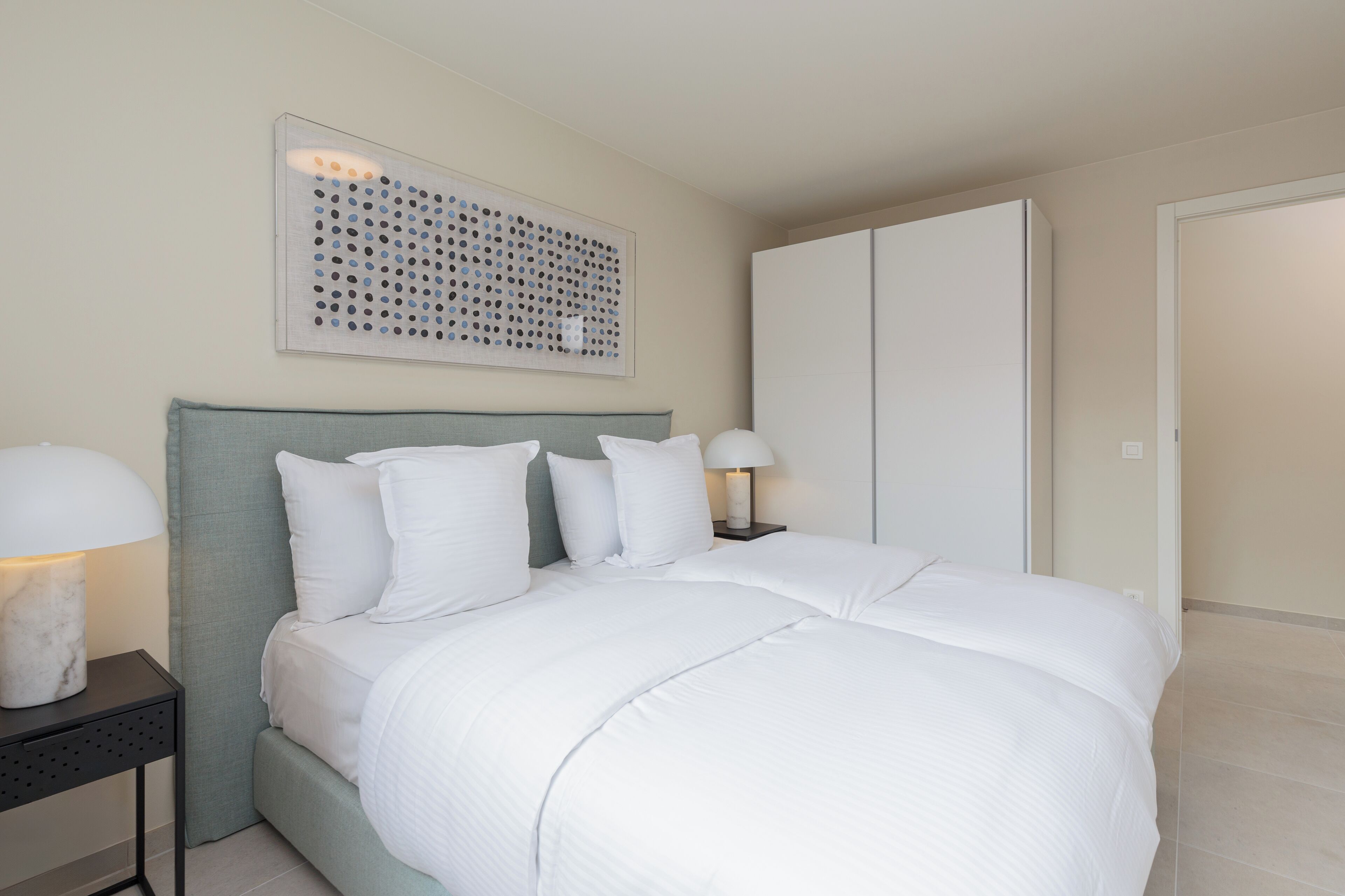A neatly made bed with white bedding, a grey headboard, and a wall-mounted artwork.