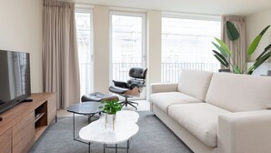 Ein modernes Wohnzimmer mit Fernseher, einem weißen Sofa, einem Ledersessel, einem Couchtisch und großen Fenstern mit Vorhängen.