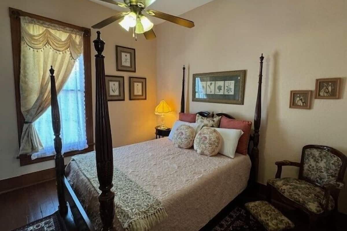 A bedroom with a four-poster bed, a ceiling fan, a chair, and framed pictures on the wall.