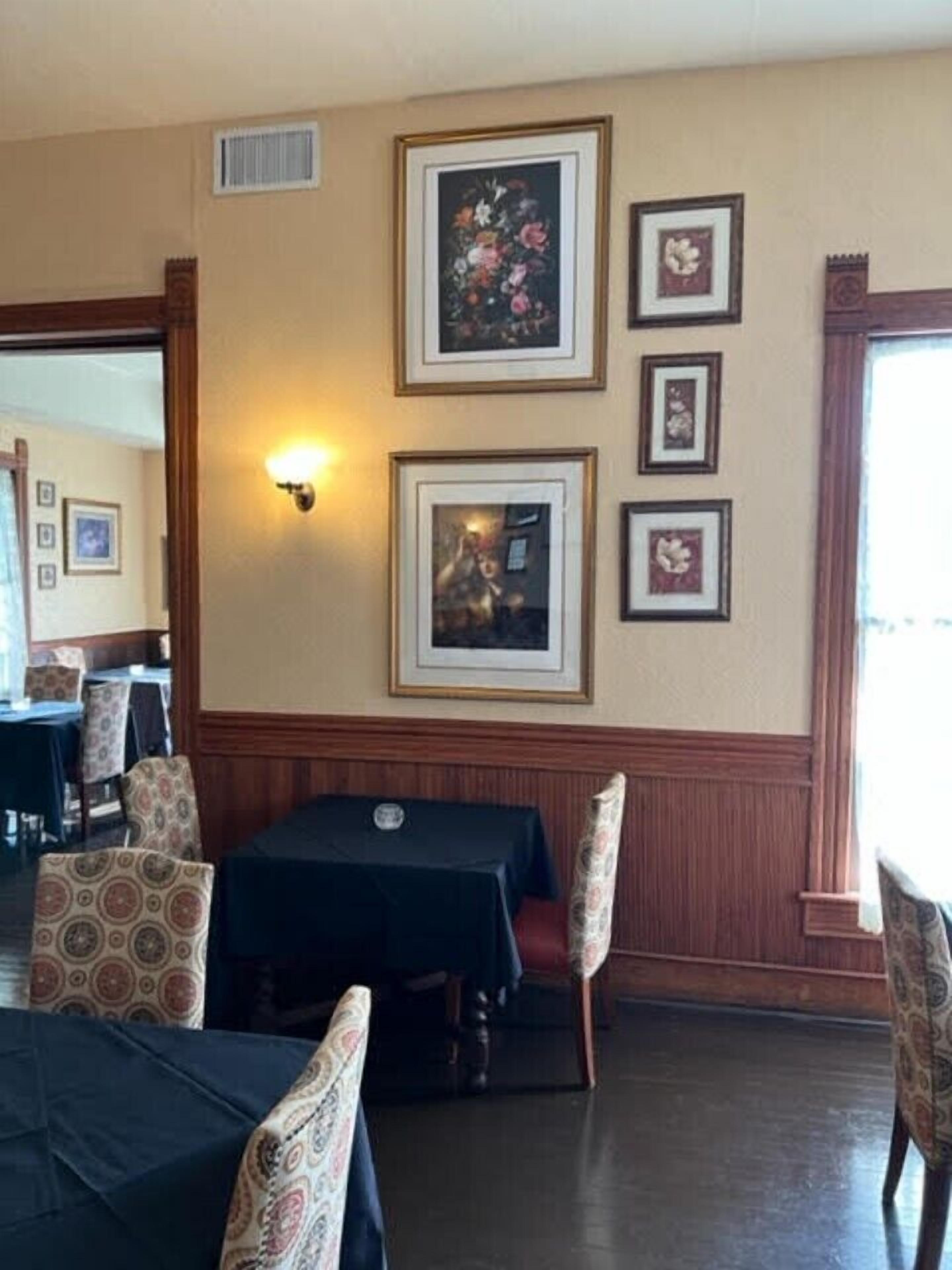 A dining area with tables and chairs, framed pictures on the wall, and a window providing natural light.