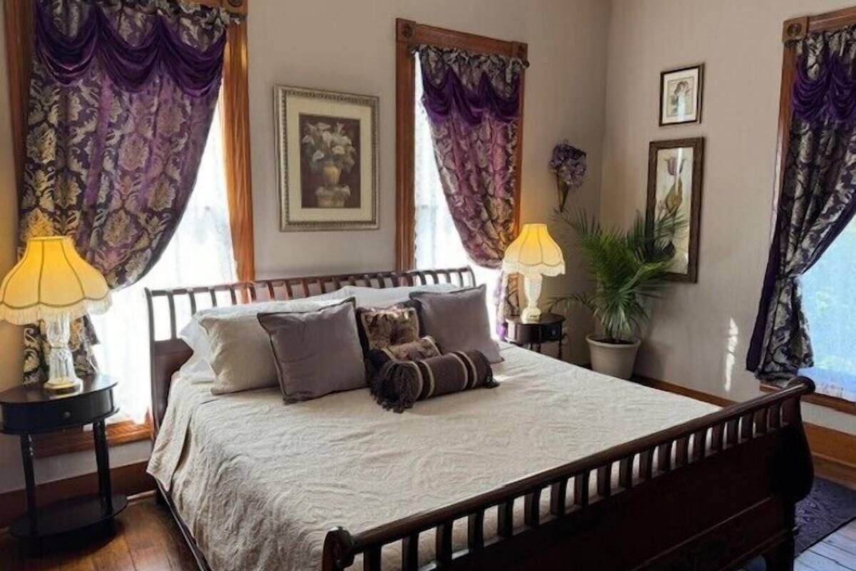 A neatly made bed with a wooden frame, two bedside tables with lamps, and a potted plant on the right.