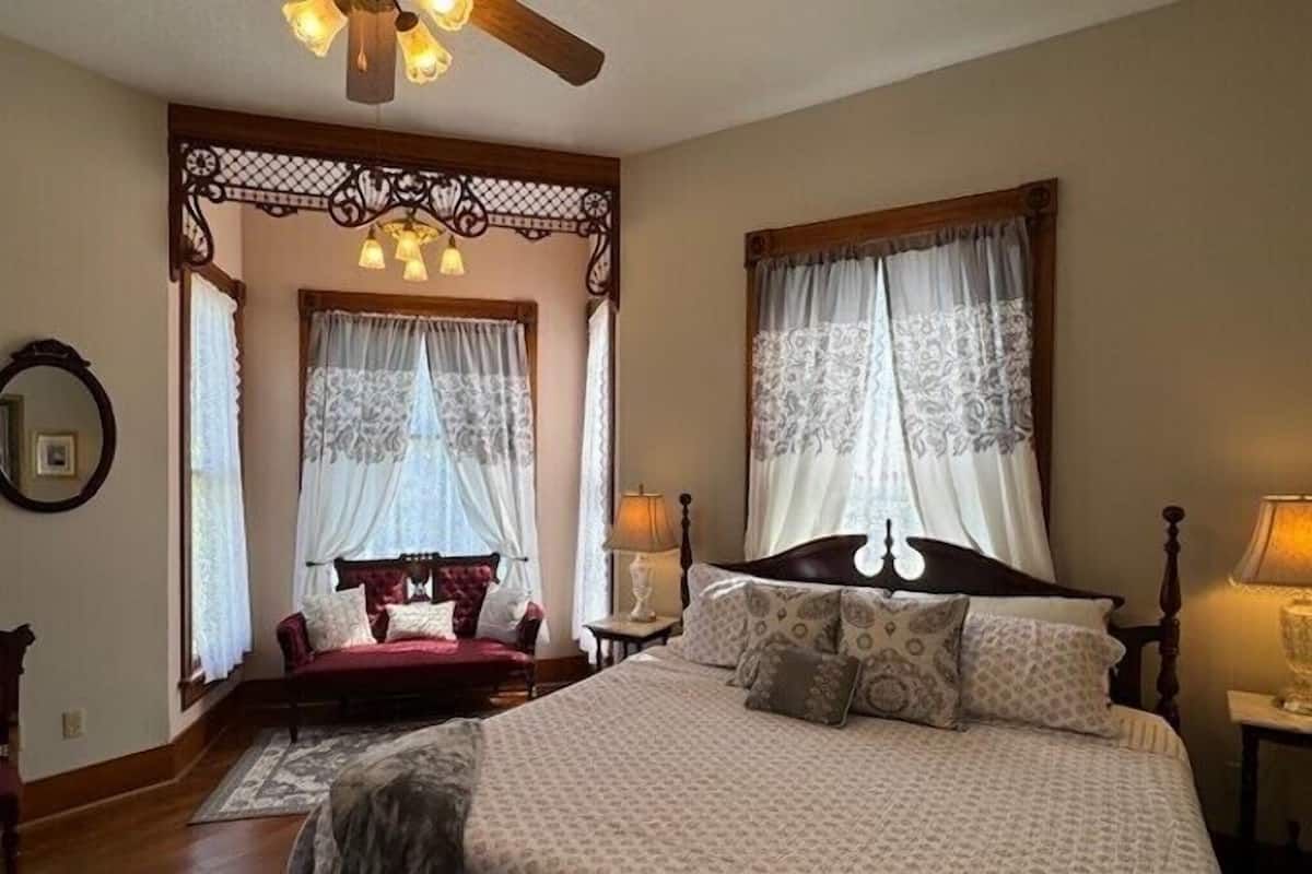 A bedroom with a large bed, two bedside tables, a ceiling fan, and a window with curtains.