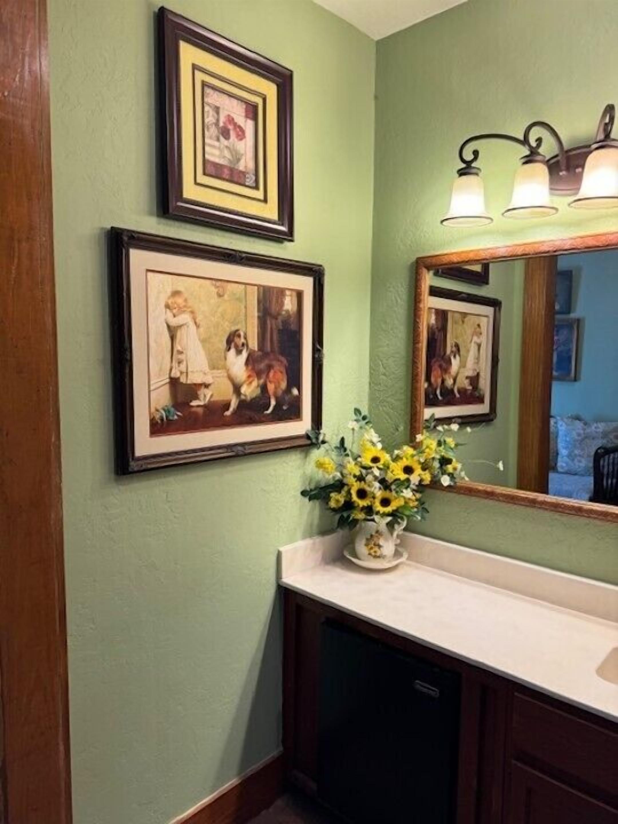 A bathroom with a green wall, framed artwork, a sink with a vase of sunflowers, and a mirror with a light fixture.