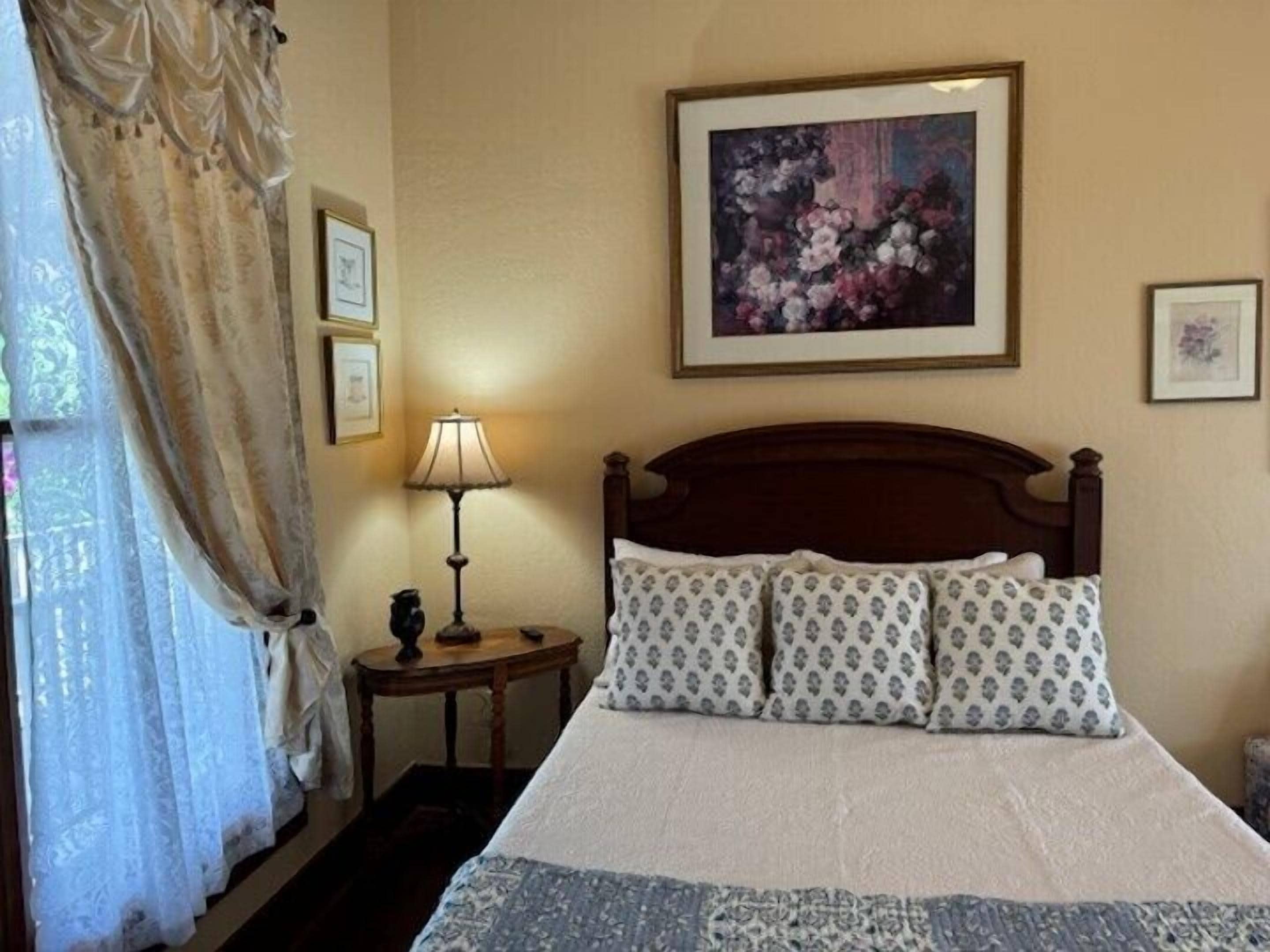 A hotel room with a large bed, a nightstand with a lamp, a framed floral painting on the wall, and a window with curtains.