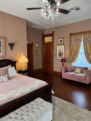 A bedroom with a bed, a chaise lounge, a chandelier, and a large window with curtains.