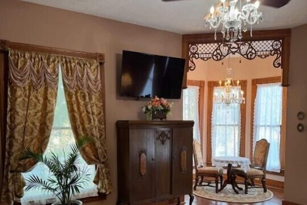 A spacious room with hardwood flooring, a large wooden cabinet, a television mounted on the wall, a chandelier, and a bed with a pink bedspread.