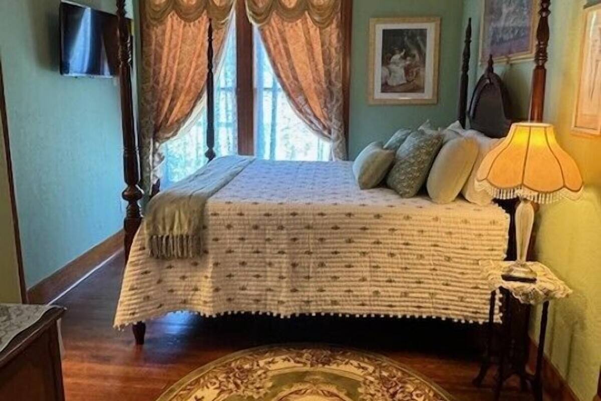 A bedroom with a bed, a ceiling fan, a window with curtains, a lamp, and a rug.