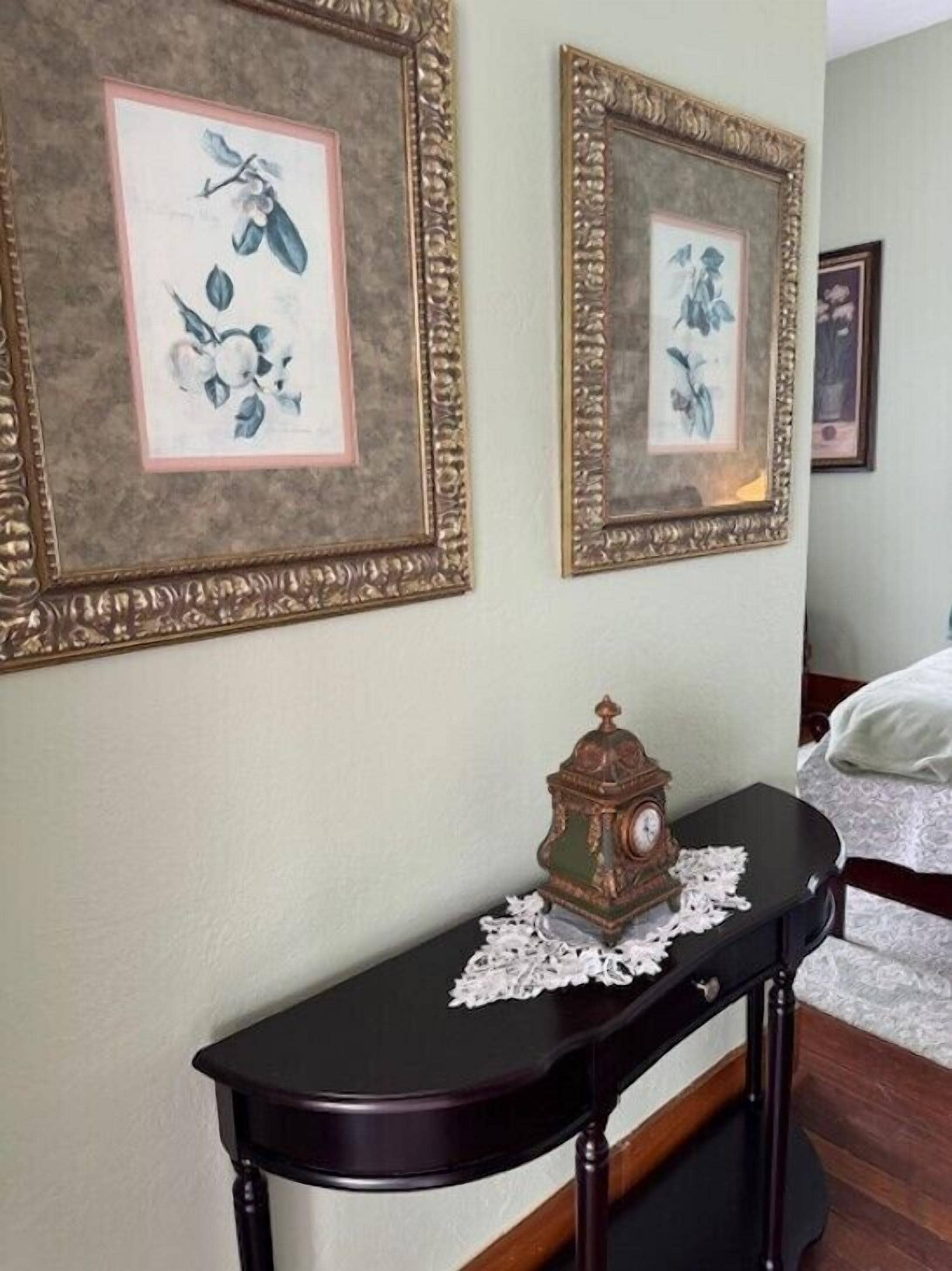 Une chambre à coucher avec deux tableaux floraux encadrés accrochés au mur, une petite table sur laquelle se trouvent une horloge et une chemin de table en dentelle, et un lit recouvert de draps blancs.