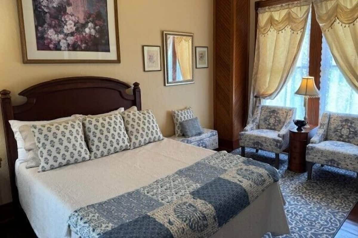 A bedroom with a large bed, two armchairs, a nightstand, and a window with curtains.