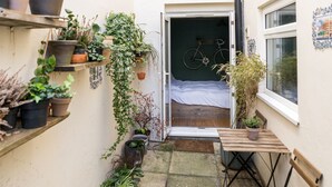 Un espacio exterior reducido con un banco de madera, una mesa pequeña y diferentes macetas con plantas.
