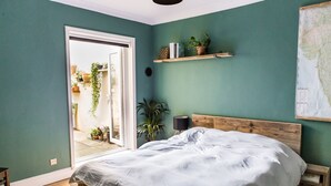 Un dormitorio con una cama, cabecera de madera, mesita de noche, lámpara y estante con libros y plantas.