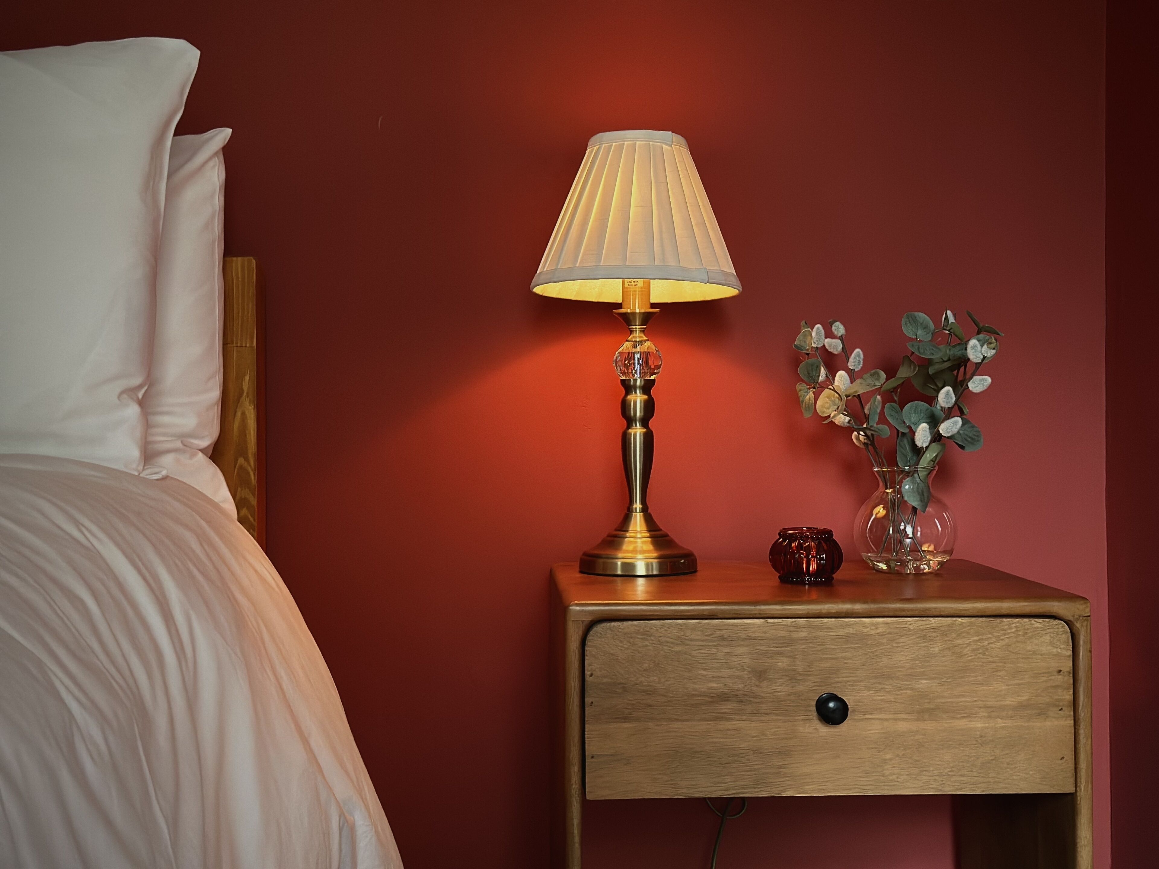 A neatly made bed with white linens, a bedside lamp with a beige shade, a small vase with green foliage, and a wooden nightstand with a drawer.