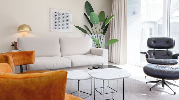 Een moderne woonkamer met een bank, twee fauteuils, een salontafel, een vloerlamp en een grote plant.