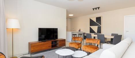 A modern living room with a flat-screen TV, a wooden cabinet, a sofa, a coffee table, and a dining area with chairs and a table.
