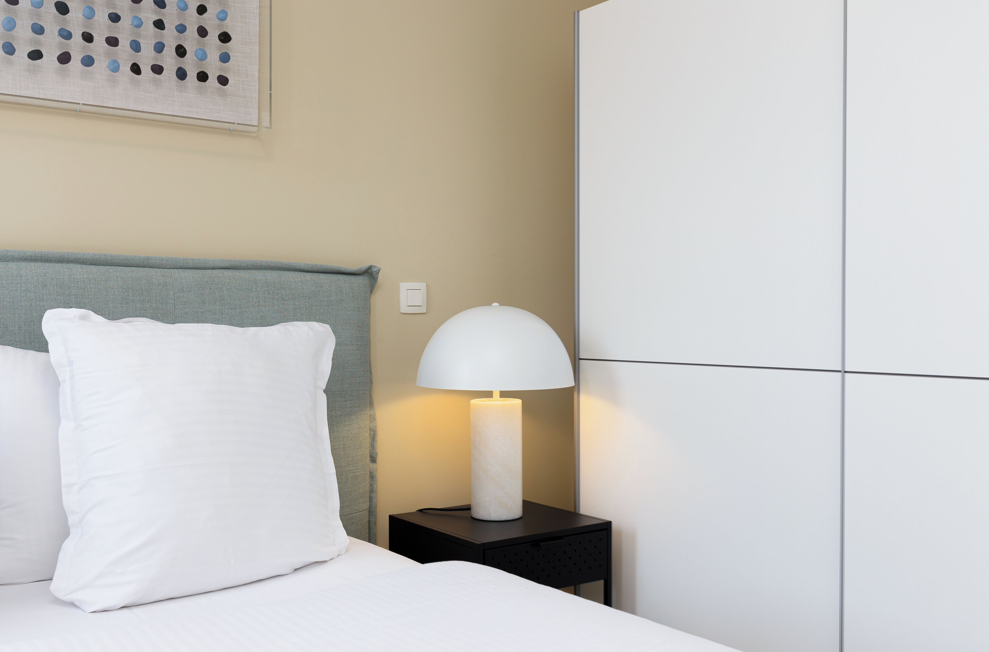Una cama bien hecha con ropa de cama y almohadas blancas, una lámpara blanca sobre una mesita de noche negra pequeña, y un armario blanco grande contra una pared color beige.