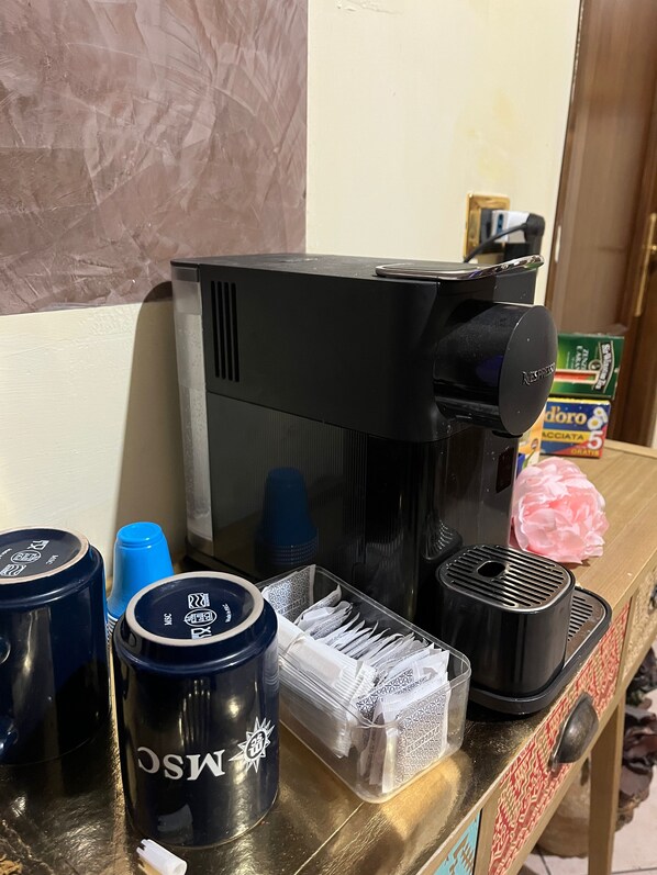 Una cafetera, una pila de tazas azules y un recipiente con sobrecitos sobre una encimera.