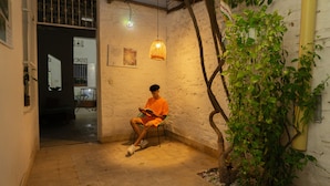 Une personne est assise sur une chaise dans une pièce dont le mur est texturé, avec un luminaire suspendu et une plante en pot.