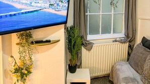 A living room with a TV displaying a cityscape, a window with plants, a white radiator, and a grey sofa. - The X Hotel (Hove)