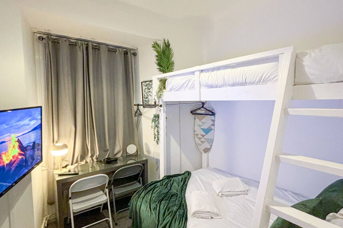 A compact room with a bunk bed, a desk with a chair, a television, and a hanging plant.