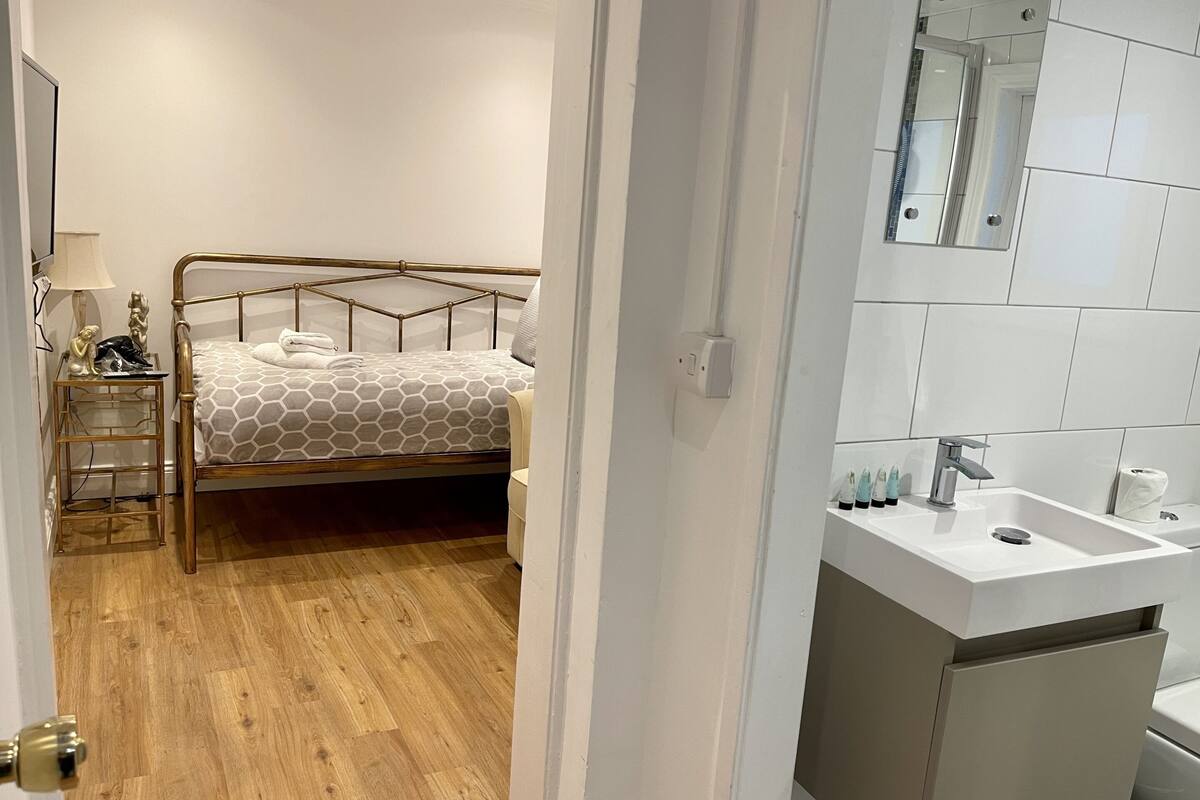 A bedroom with a metal bed frame, a bedside table with a lamp, and a bathroom with a sink and mirror.