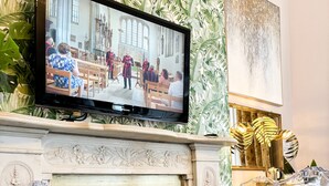 A room with a television mounted above a fireplace, clear plastic chairs around a small table, and a patterned wallpaper. - The X Hotel (Hove)