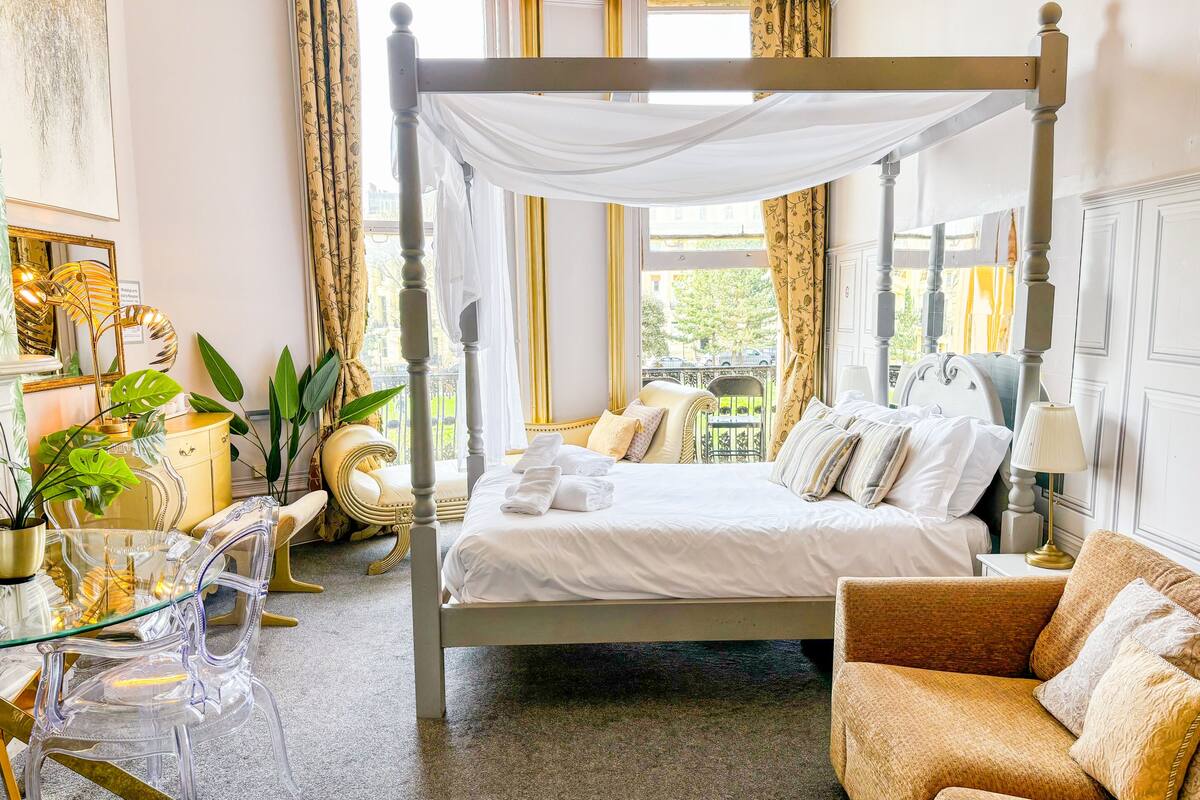 A four-poster bed with white linens, a glass coffee table, a brown sofa, and a view of the outdoors through large windows.