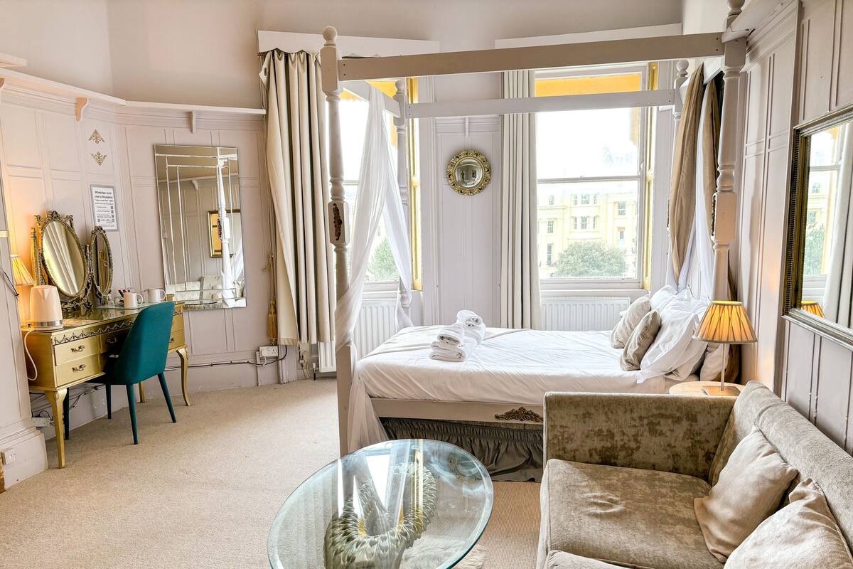 A hotel room with a bed, a glass table, a sofa, a vanity with mirrors, and a window with curtains.