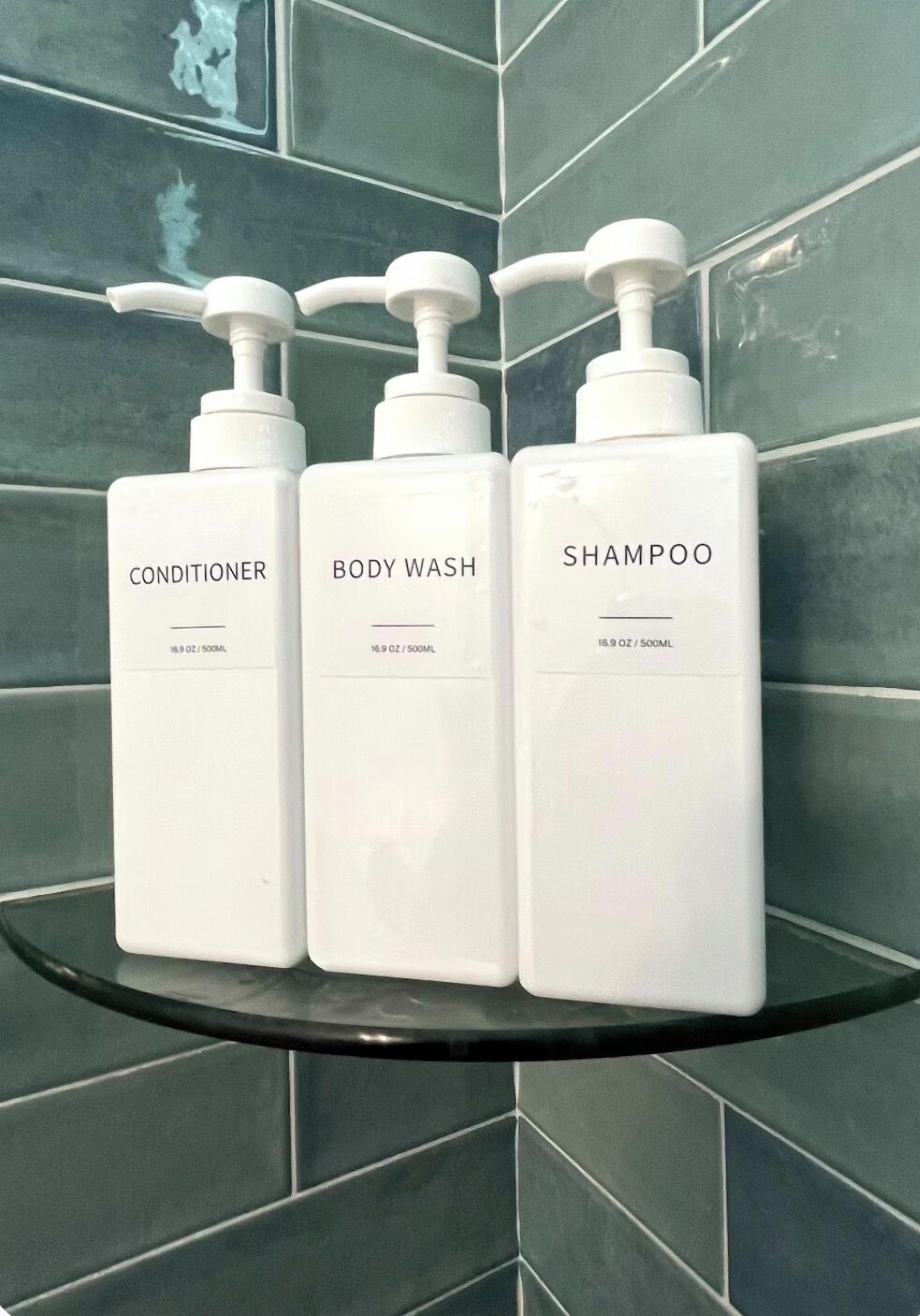Kolme valkoista pulloa, joissa on hoitoainetta, vartalopesua ja shampoota, mustalla hyllyllä laatoitetun seinän edessä.