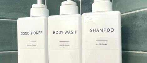 Kolme valkoista pulloa, joissa on hoitoainetta, vartalopesua ja shampoota, mustalla hyllyllä laatoitetun seinän edessä.