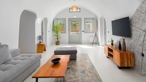 A modern living room with a grey sofa, a wooden coffee table, a grey ottoman, a wooden TV stand, and a large TV mounted on a stone wall. - White Occhio Villa Santorini (Santorini)