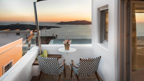 Un balcón con vista al mar al atardecer, con dos sillas con cojines estampados y una pequeña mesa con una bebida.