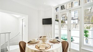 A dining area with a round table set for four, a flat-screen TV mounted on the wall, and a view of a cityscape through large glass doors. - 4 bedroom home by Buckingham Palace (London)